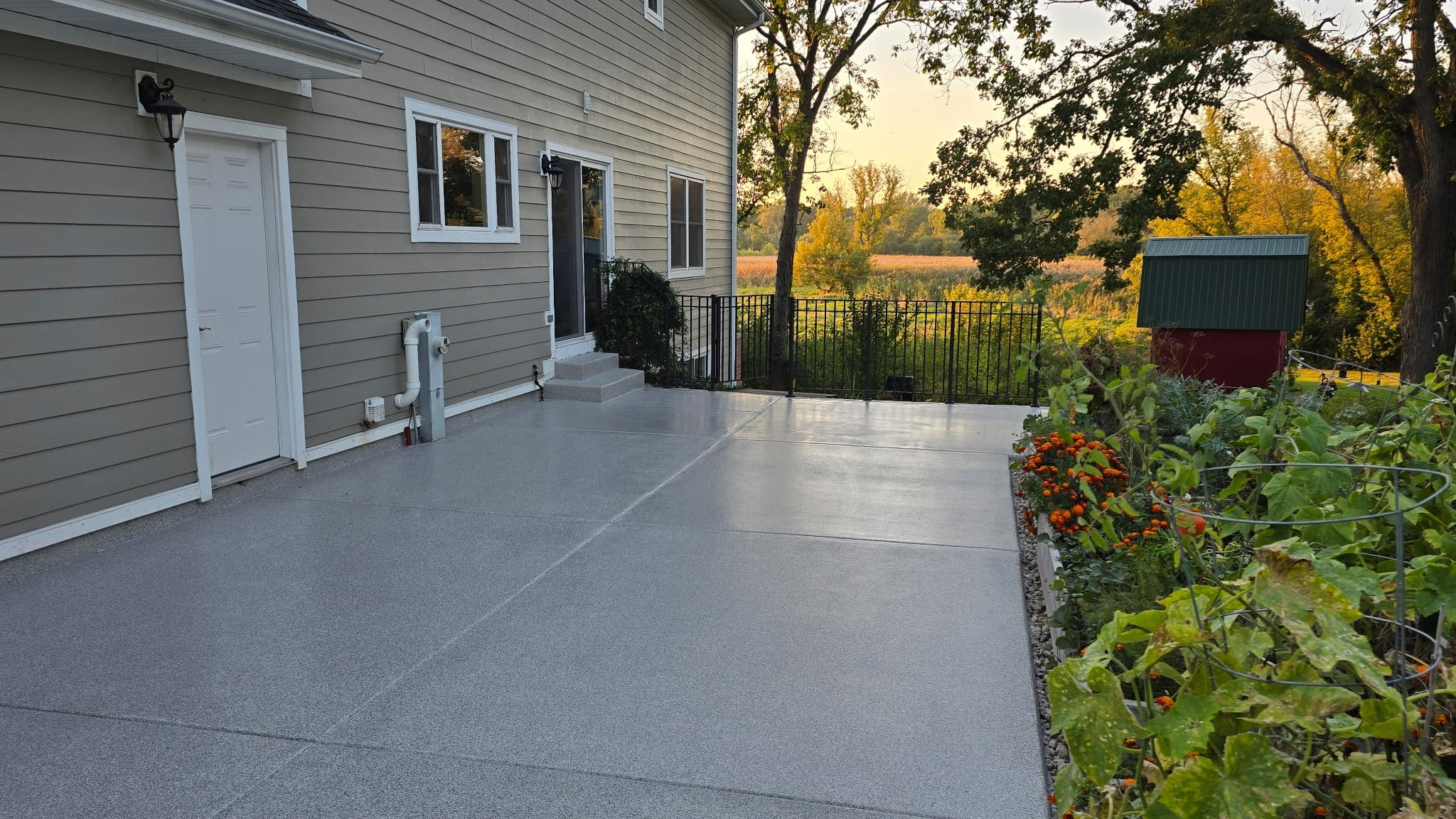 Coated backyard patio in grey flake running alongside a home at sunset, with a garden bed and open fields beyond