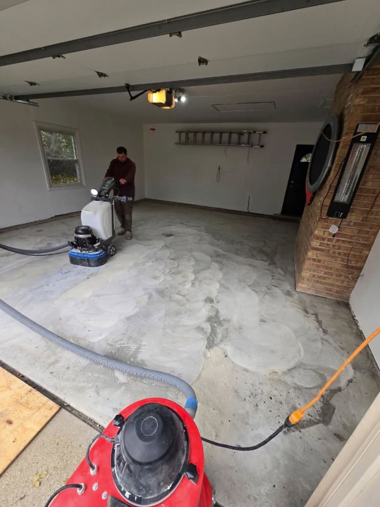 Technician running a vacuum-shrouded diamond grinder across a garage slab, cutting the swirl profile the coating bonds into